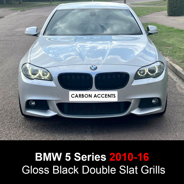 A silver BMW 5 Series (2010-16) with gloss black double slat grills is parked on a street, surrounded by grass. The front plate reads "CARBON ACCENTS."