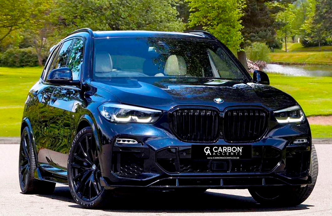 A sleek, black SUV with illuminated headlights is parked on a paved surface in a green park setting. The license plate reads, "CARBON ACCENTS www.carbonaccents.co.uk."