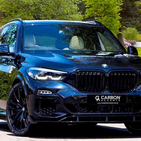 A sleek, black SUV with illuminated headlights is parked on a paved surface in a green park setting. The license plate reads, "CARBON ACCENTS www.carbonaccents.co.uk."