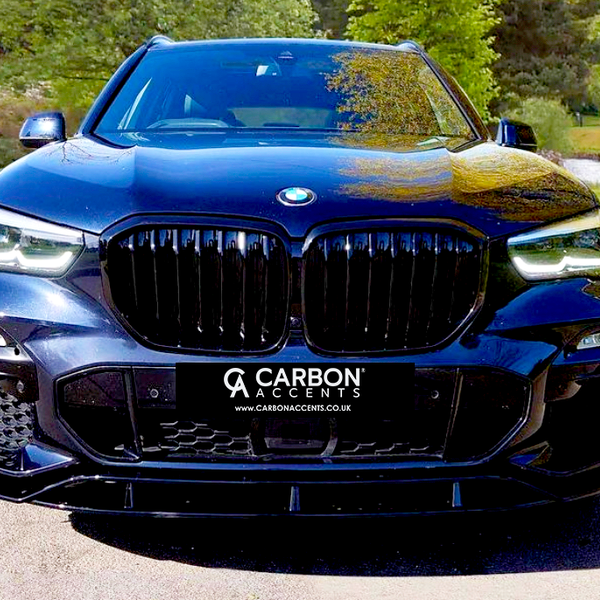 A black SUV is parked facing forward, showcasing its sleek design and prominent grille. Surrounding it are green trees and a pond. Text on the car reads, "CARBON ACCENTS www.CARBONACCENTS.co.uk".