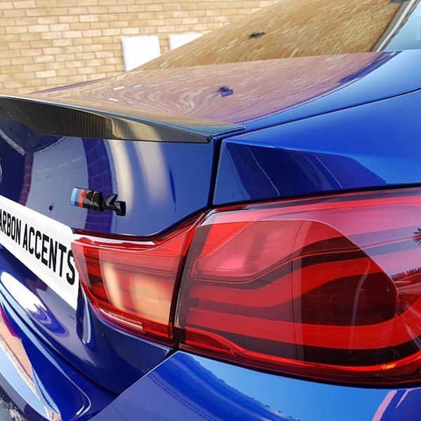 A blue BMW car with a carbon fiber spoiler is parked near a brick wall. The license plate reads "CARBON ACCENTS," and there's an "M4" badge visible.