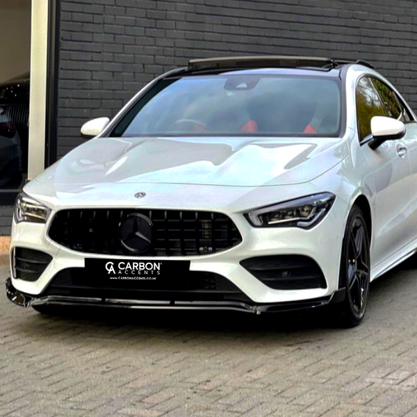 A sleek white car is parked on a brick driveway, with a black grille and wheels, beside a dark gray building. The license plate reads “CARBON ACCENTS www.carbonaccents.co.uk.”