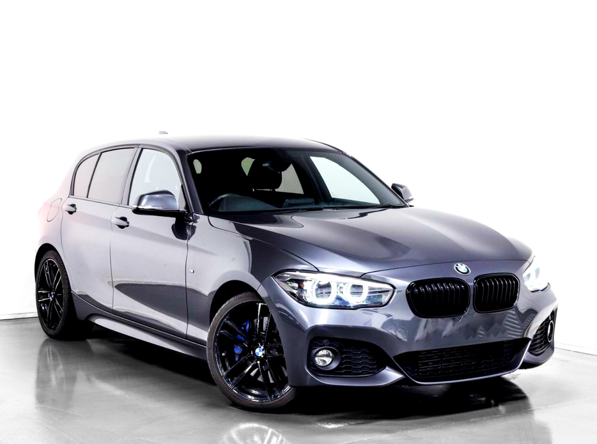 A gray BMW hatchback, featuring sleek black alloy wheels and prominent headlights, is parked on a reflective gray floor against a white background.