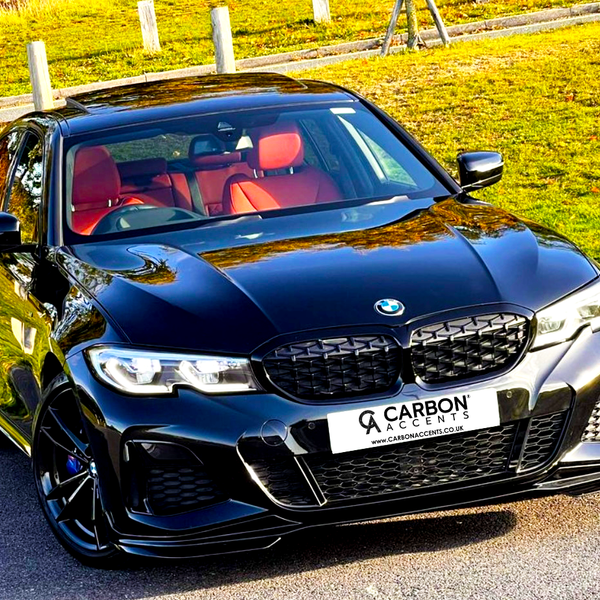 A black BMW sedan with red interior is parked on a paved road, surrounded by grassy areas. The license plate reads "CARBON ACCENTS, WWW.CARBONACCENTS.CO.UK."