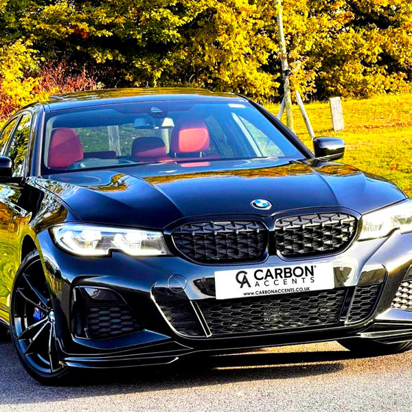 A black car with red seats is parked on a paved surface. Behind it, trees display autumn colors. The license plate reads: "CARBON ACCENTS www.CARBONACCENTS.CO.UK".