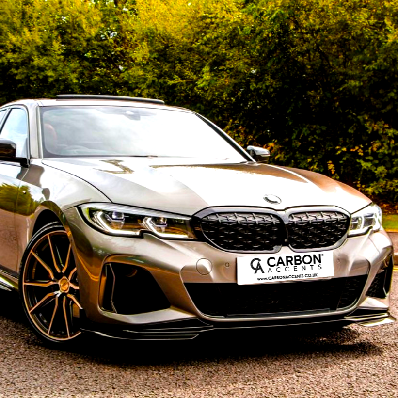 A sleek, silver car is parked on a road. The license plate reads "CARBON ACCENTS www.carbonaccents.co.uk." Behind, lush green trees create a vibrant, natural backdrop.