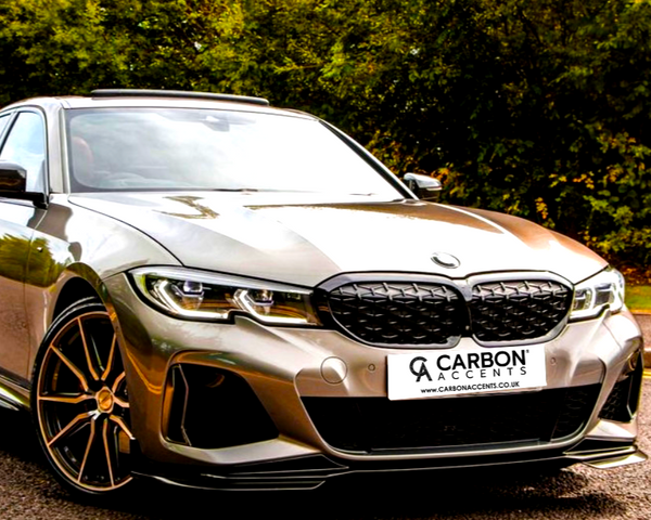 A sleek, silver car is parked on a road. The license plate reads "CARBON ACCENTS www.carbonaccents.co.uk." Behind, lush green trees create a vibrant, natural backdrop.