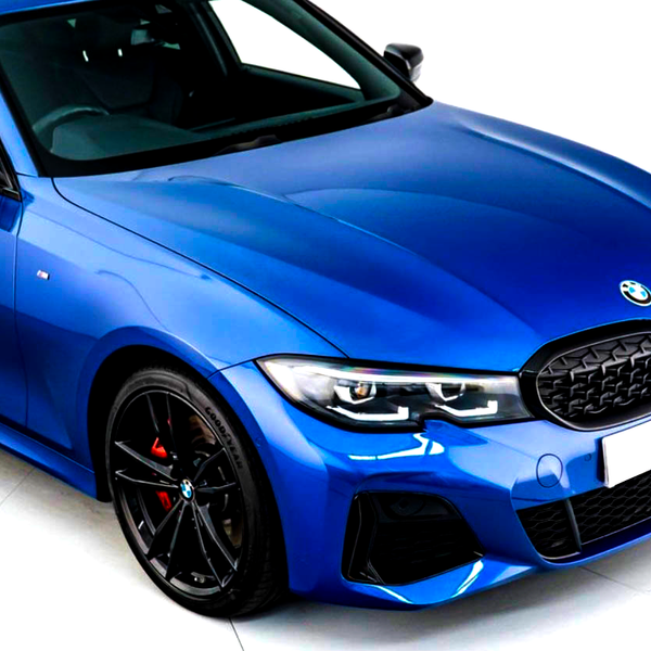 A metallic blue BMW car is stationary indoors, featuring a black grille, sporty headlights, and black wheels with red brake calipers. The environment is brightly lit, enhancing its sleek design.
