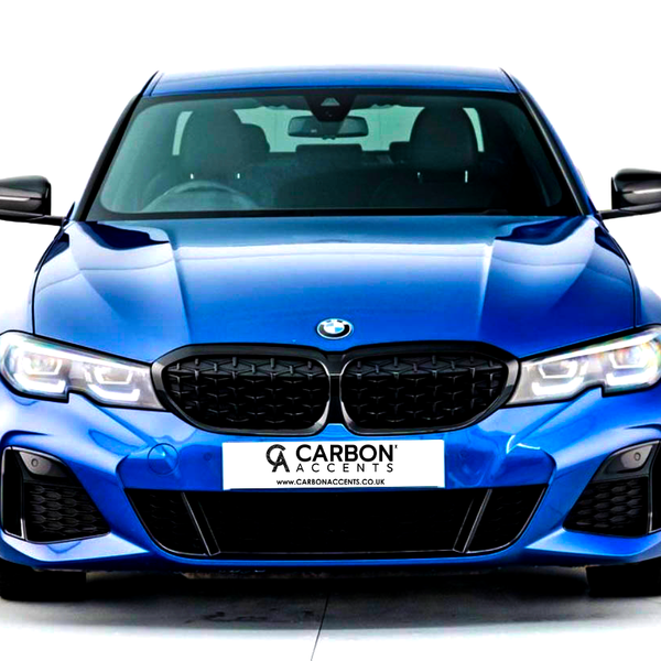 A blue BMW car is parked facing forward, with illuminated headlights. The background is white. The license plate reads: "CARBON ACCENTS" with the URL "www.carbonaccents.co.uk."