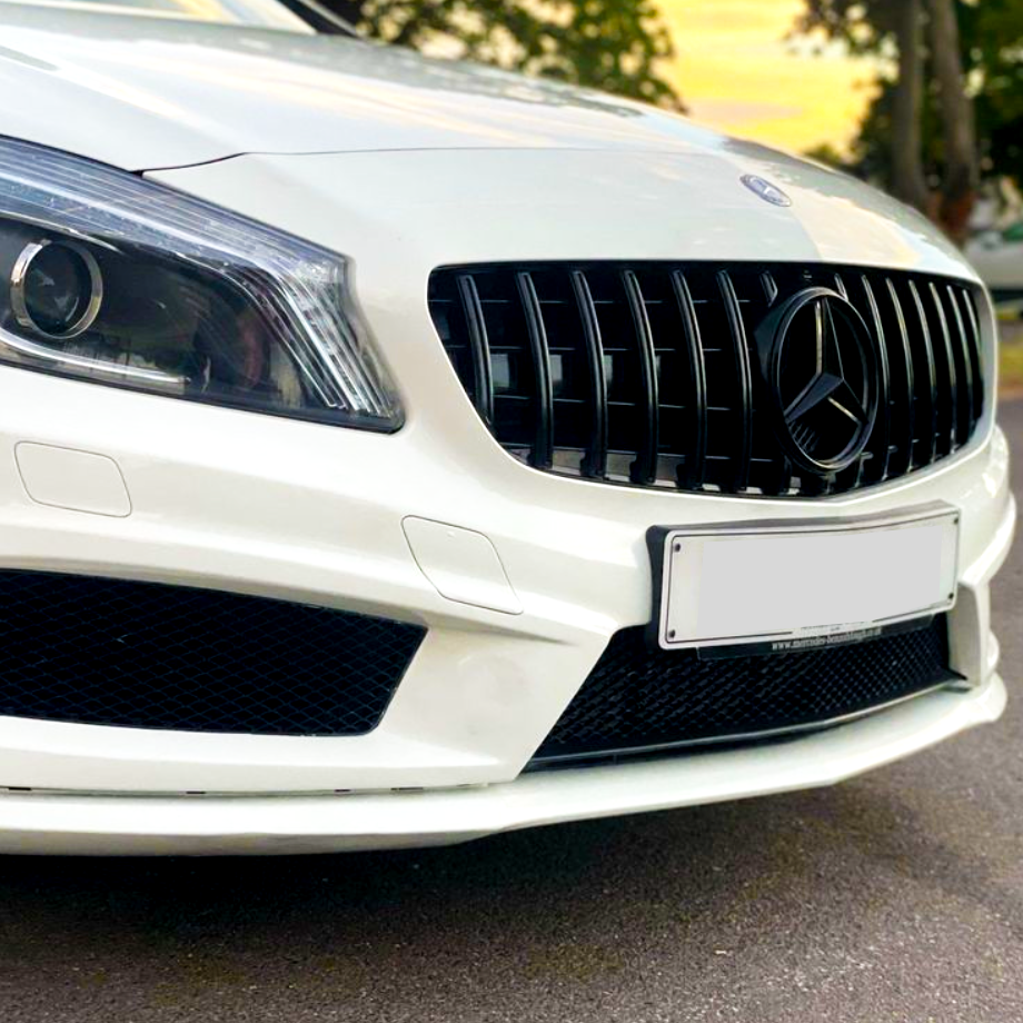 A white car's front view prominently displays a black grille and headlight, parked on a road with blurred greenery and a sunset sky in the background. There is no visible text.
