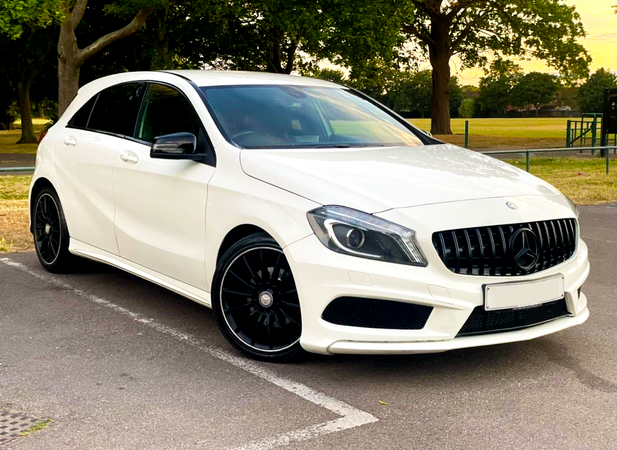 A white Mercedes-Benz hatchback car is parked on a paved area, surrounded by trees and grassland, under a partly cloudy sky.