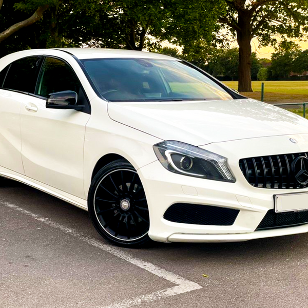 A white Mercedes-Benz hatchback car is parked on a paved area, surrounded by trees and grassland, under a partly cloudy sky.