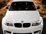 A white BMW car, parked on a rural road, is surrounded by grass and trees with sunlight casting shadows.