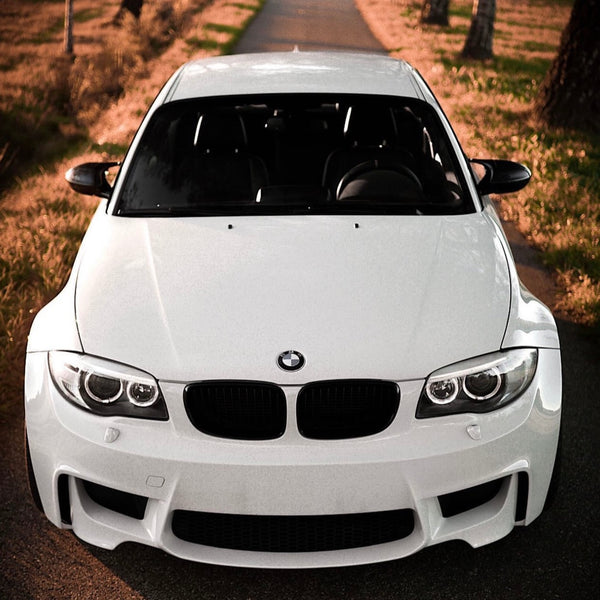 A white BMW car, parked on a rural road, is surrounded by grass and trees with sunlight casting shadows.
