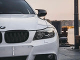 A white BMW car is parked, partially shown from the front. It features a sleek grille and headlights, located in a waterfront area with a calm atmosphere.