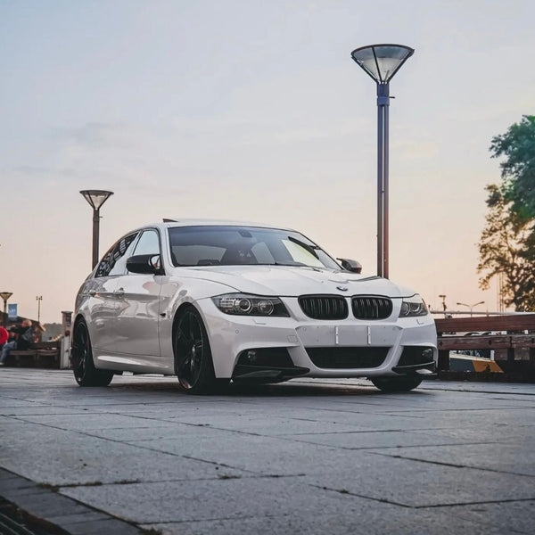 A white car is parked on a paved area, surrounded by street lamps and trees against a backdrop of a clear sky.