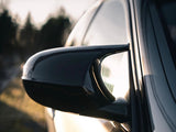 Car side mirror reflects sunlight, with a sleek black finish. The vehicle is parked outdoors, and the background features blurred silhouettes of trees under a bright sky.