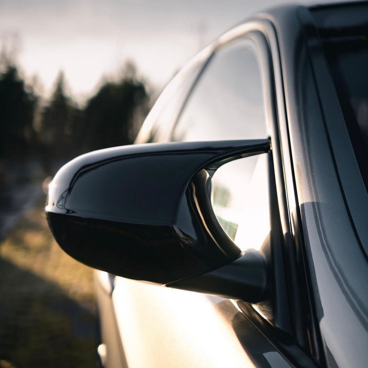 Car side mirror reflects sunlight, with a sleek black finish. The vehicle is parked outdoors, and the background features blurred silhouettes of trees under a bright sky.