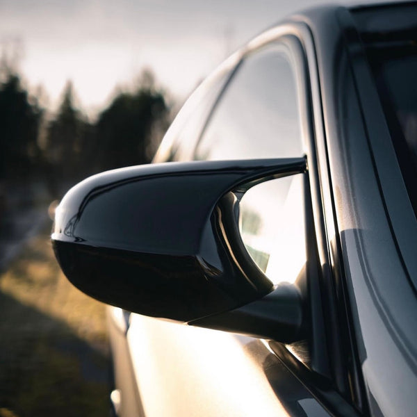 Car side mirror reflects sunlight, with a sleek black finish. The vehicle is parked outdoors, and the background features blurred silhouettes of trees under a bright sky.