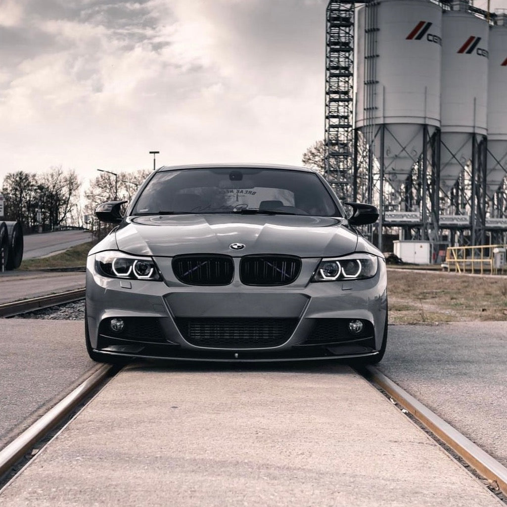 A gray BMW car stands still, positioned on train tracks. In the background, large industrial silos rise against a cloudy sky.