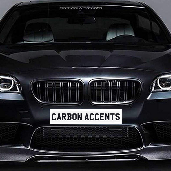 The car is a sleek black sedan with illuminated headlights facing forward. Text on the front reads "CARBON ACCENTS." It's positioned in a dimly lit studio environment.