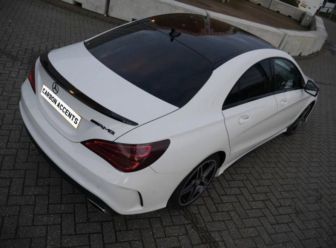 A white car parked on a brick surface features a black rooftop and spoiler. The rear badge reads "AMG" and the license plate text reads "CARBON ACCENTS." Concrete barriers are in the background.
