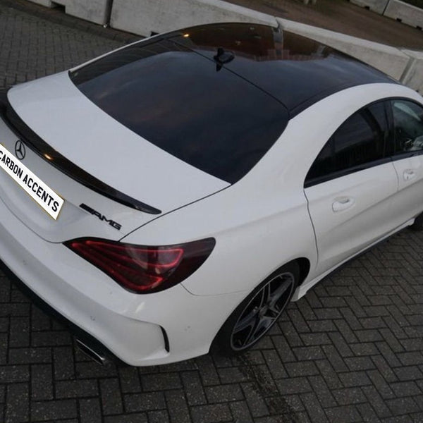 A white car parked on a brick surface features a black rooftop and spoiler. The rear badge reads "AMG" and the license plate text reads "CARBON ACCENTS." Concrete barriers are in the background.