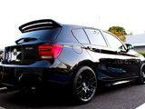 A black BMW 135i parked in a lot, featuring tinted windows, a rear spoiler, and glossy finish. Surrounding context includes greenery and a building.