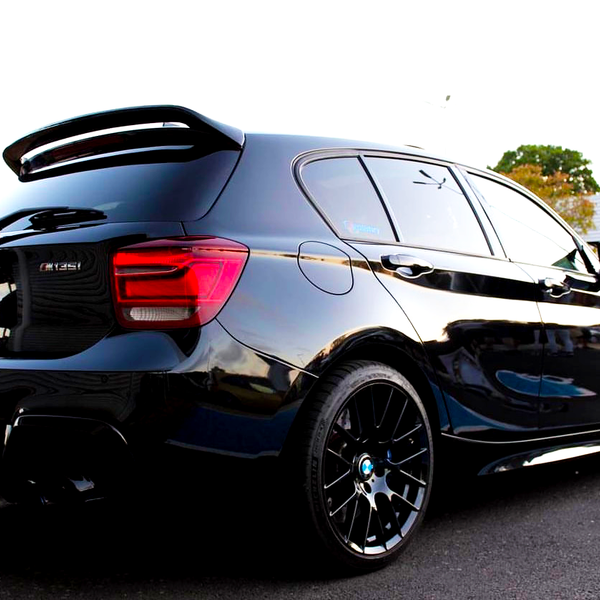 A black BMW 135i parked in a lot, featuring tinted windows, a rear spoiler, and glossy finish. Surrounding context includes greenery and a building.