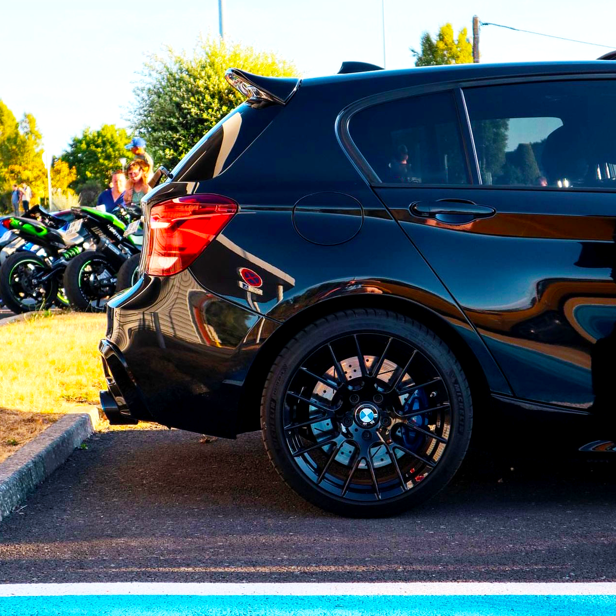 A black car with custom rims is parked beside a blue line in a lot. In the background, several motorcycles are parked near green bushes and a pole.