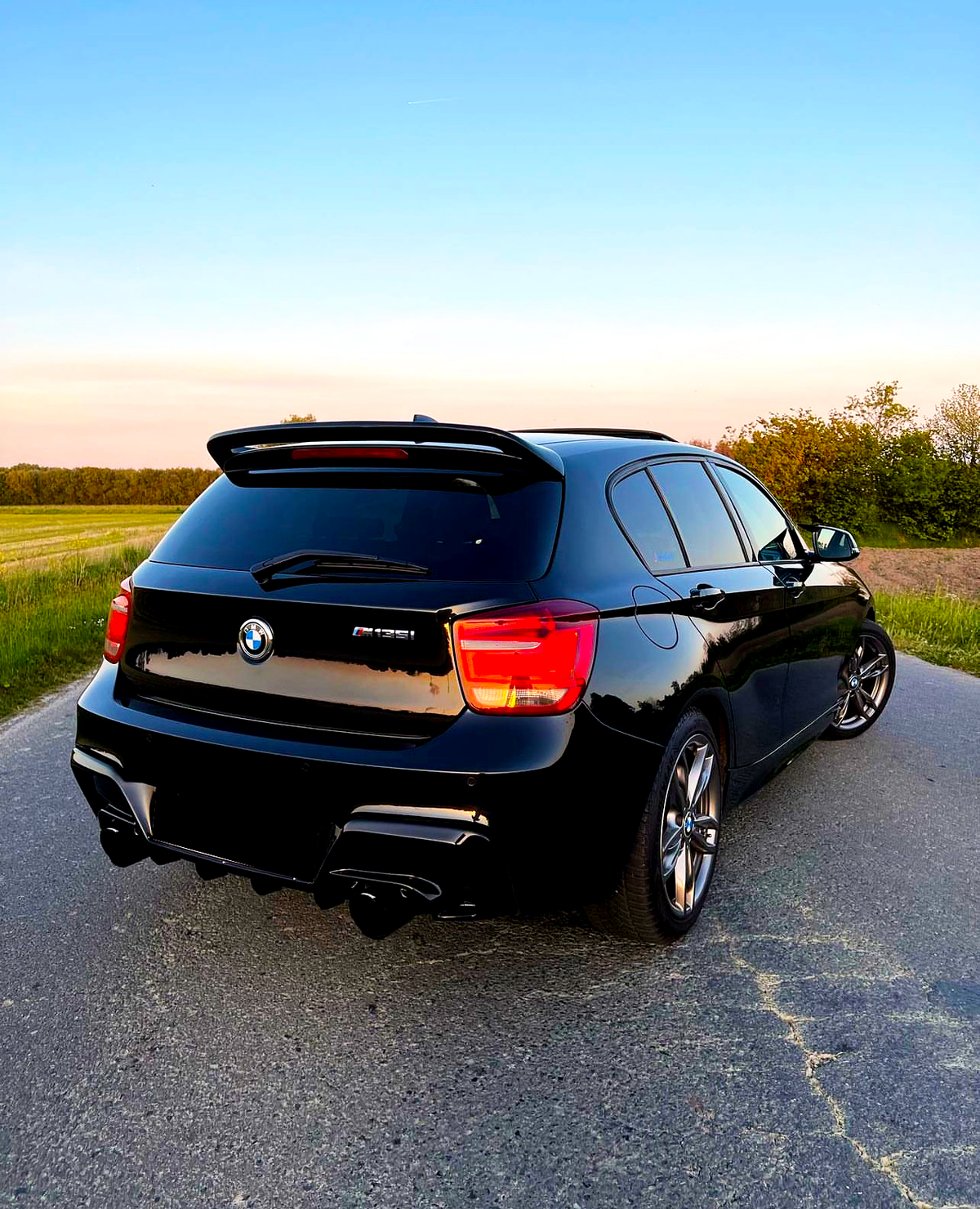 A black BMW M135i is parked on a deserted rural road during sunset, surrounded by green grass and trees under a clear blue sky.