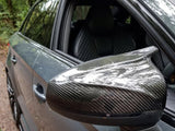 A carbon fiber side mirror on a sleek car reflects the forest canopy above, with partially visible quilted leather seats inside the vehicle. The car is parked on a forest path.