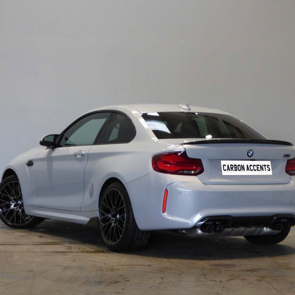 A silver BMW coupe parked indoors on a concrete floor, featuring black wheels and a visible “CARBON ACCENTS” sticker on its rear.