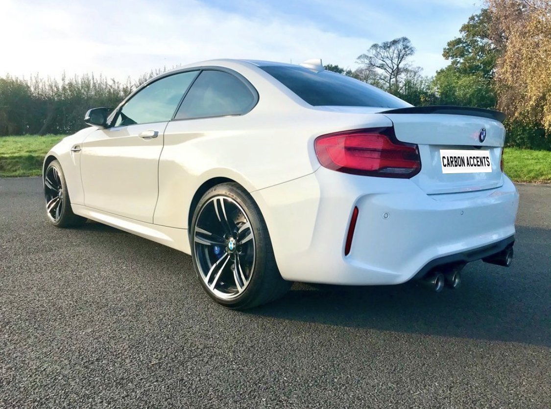 A white sports car is parked on an asphalt road. It features tinted windows, sleek design, and dual exhausts. License plate reads "CARBON ACCENTS." Background shows trees and a grassy field.