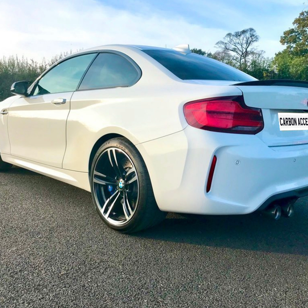 A white sports car is parked on an asphalt road. It features tinted windows, sleek design, and dual exhausts. License plate reads "CARBON ACCENTS." Background shows trees and a grassy field.