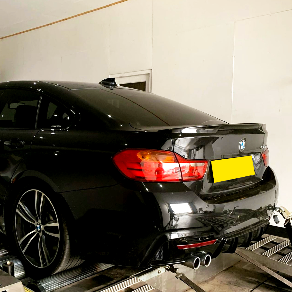 A black car is secured on a testing rig in a white-walled room. The car has red tail lights illuminated and dual exhaust pipes. A strap is attached to its rear.