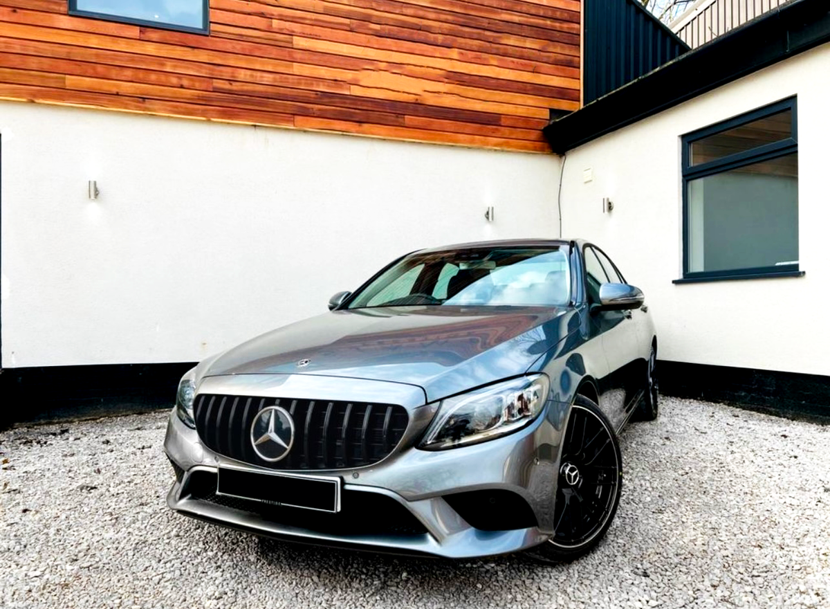 A silver car is parked on gravel, featuring a prominent grille with a star emblem. It's positioned near a modern building with white and wood-paneled walls.