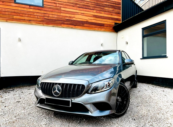 A silver car is parked on gravel, featuring a prominent grille with a star emblem. It's positioned near a modern building with white and wood-paneled walls.
