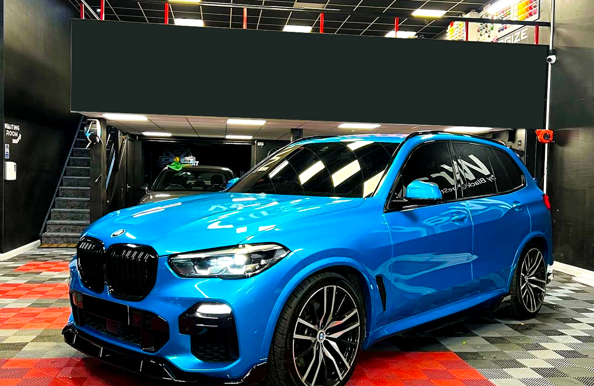 A blue BMW SUV is parked in a garage with a black ceiling and checkered red and black floor. "NAV" is written on the driver's side window.