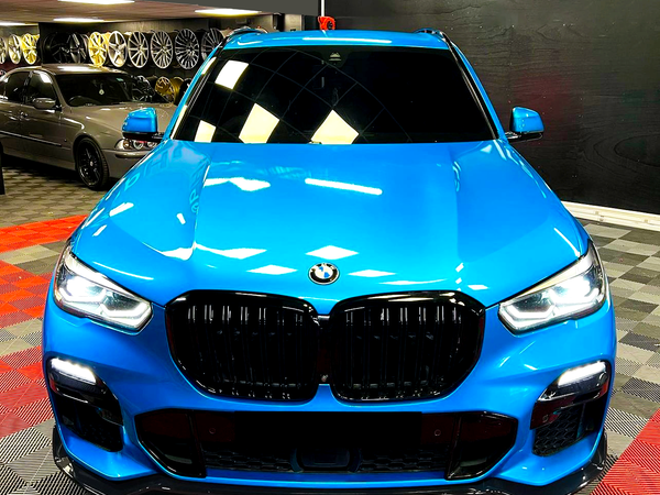 A blue BMW SUV is parked with headlights on in a showroom, surrounded by various wheels displayed on the wall and a checkered floor pattern.