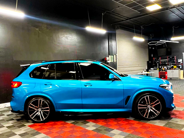 A bright blue SUV is parked in a garage with black walls and a checkered floor, under fluorescent lights, near a rolling door and shelves with tools.