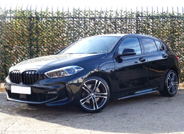 A black car is parked on a paved surface, with a metal fence covered in foliage behind it. The car features stylish alloy wheels and glossy paint reflecting sunlight.