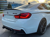 A white BMW M440i is parked on a city street at sunset, featuring illuminated red taillights and dual exhaust pipes, adjacent to modern buildings.