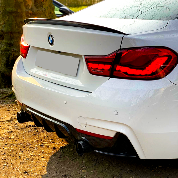 A white BMW car, parked on a dirt surface, has its red taillights illuminated. The rear bumper and dual exhausts are visible, with a shadow cast in the golden hour sunlight.
