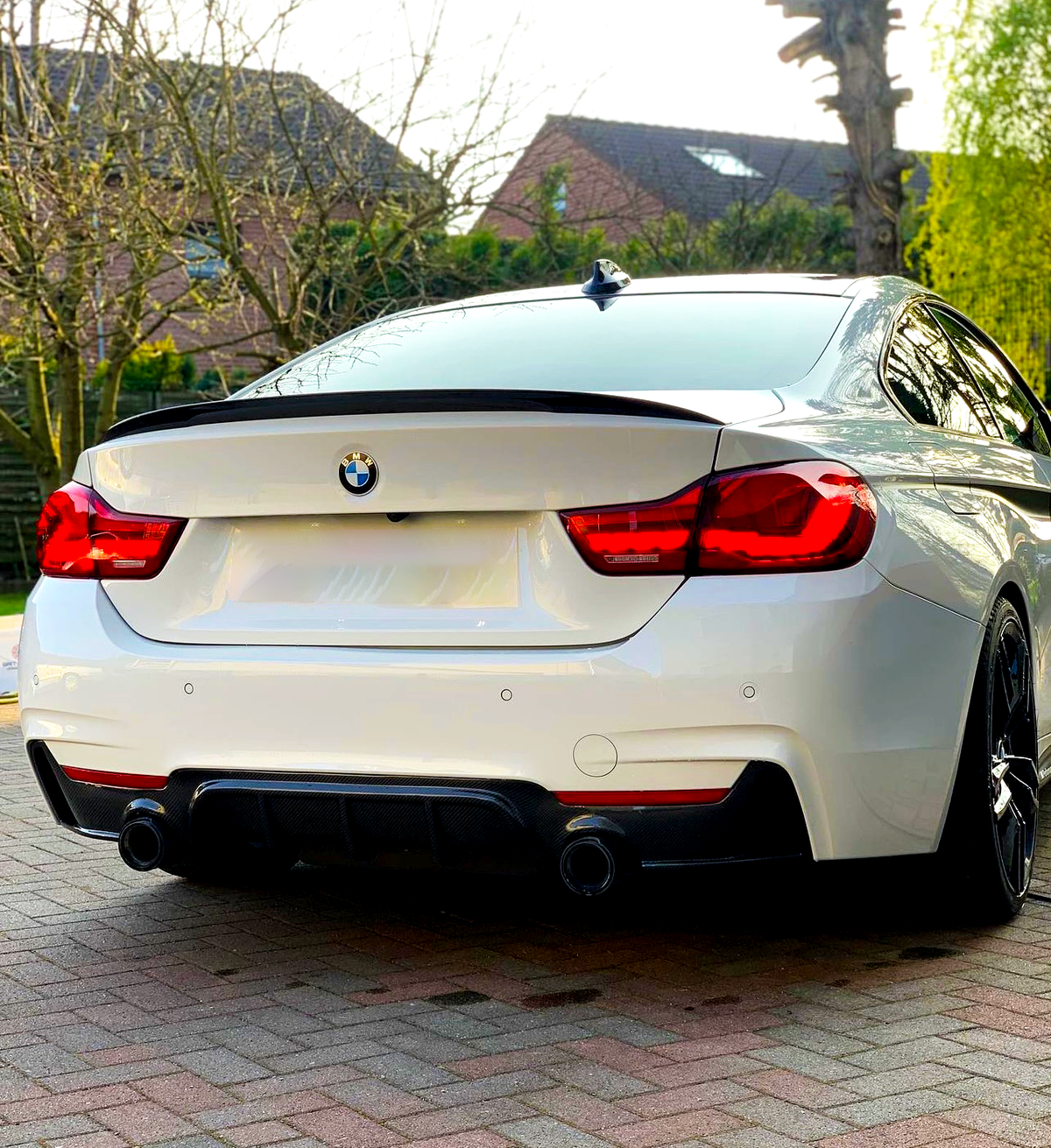 A white BMW car is parked on a paved driveway. It's surrounded by trees and brick houses in the background, with the sun casting a warm glow.