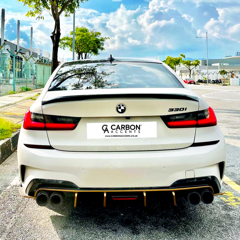 A white BMW 330i is parked on a sunny street. It features dual exhausts and displays the "CARBON ACCENTS" license plate with "www.CARBONACCENTS.CO.UK." Urban buildings and trees are visible.