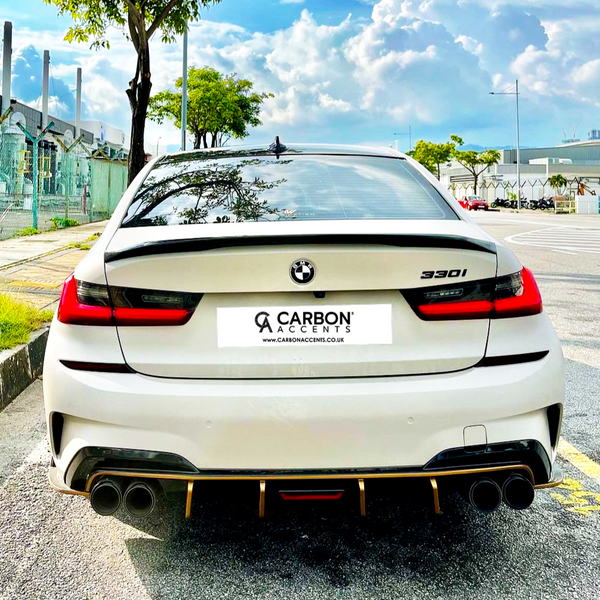 A white BMW 330i is parked on a sunny street. It features dual exhausts and displays the "CARBON ACCENTS" license plate with "www.CARBONACCENTS.CO.UK." Urban buildings and trees are visible.
