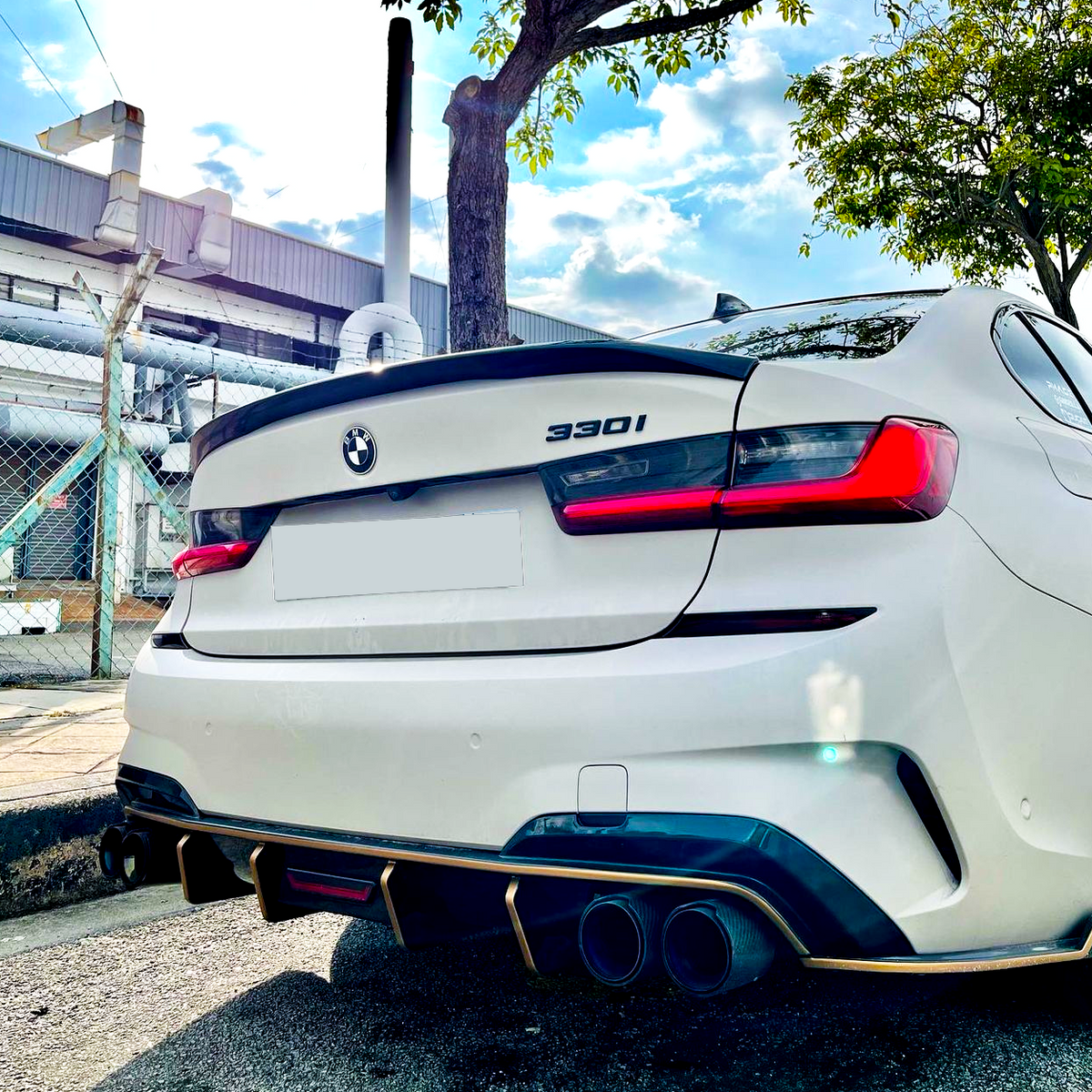 A white BMW 330i car is parked on a street, with visible dual exhaust pipes and a factory building and trees in the background under a blue sky.