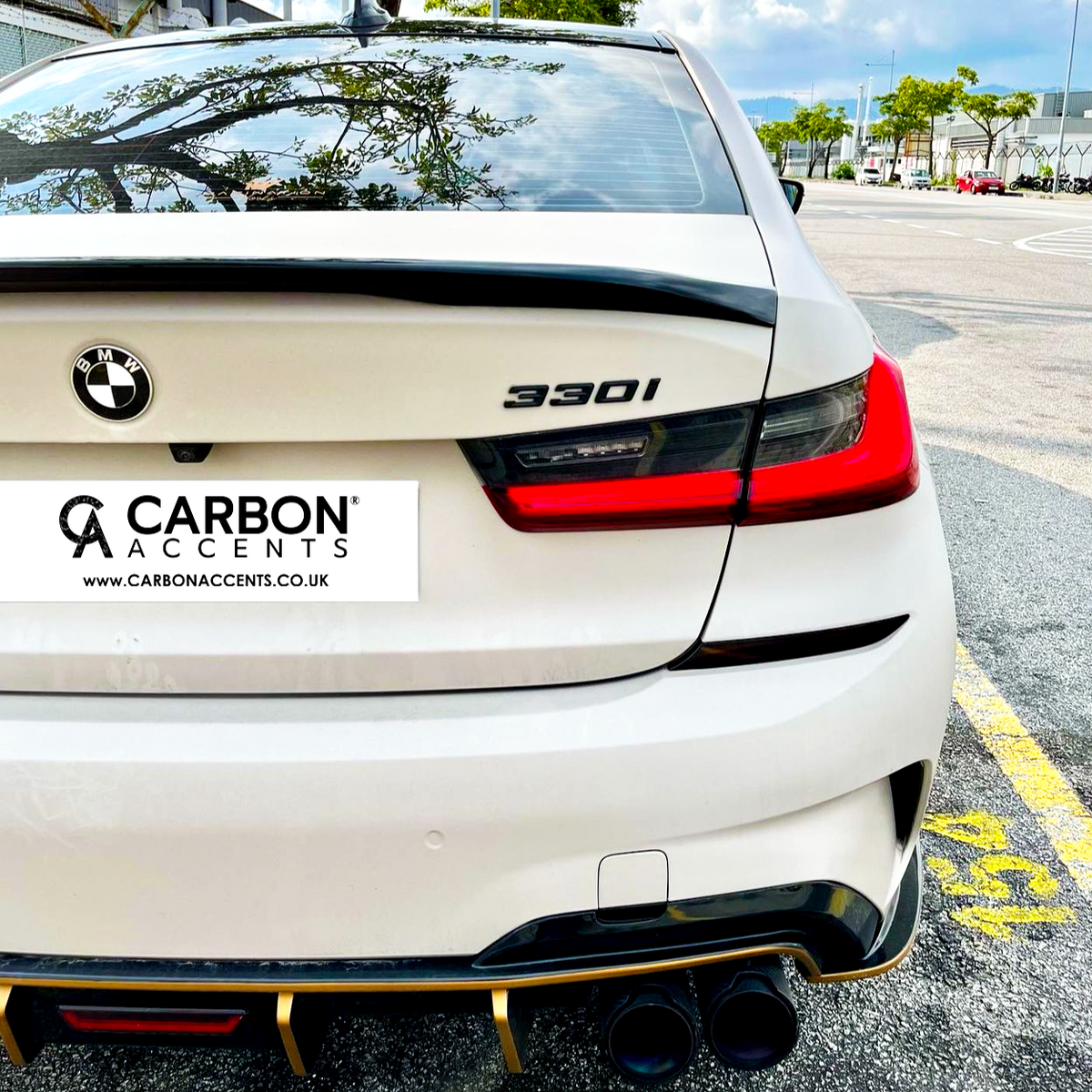 BMW car rear showcasing "330i" badge and dual exhausts; parked in a lot with trees and buildings visible. Text: “CARBON ACCENTS” and “www.CARBONACCENTS.CO.UK”.