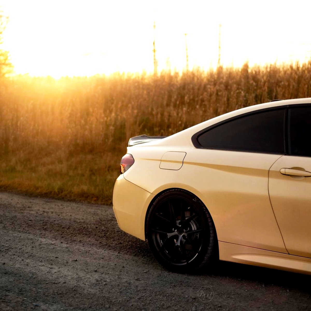 A yellow car is parked on a gravel road, with golden sunlight illuminating a field of tall grass in the background. The car's back wheel and tinted windows are visible.